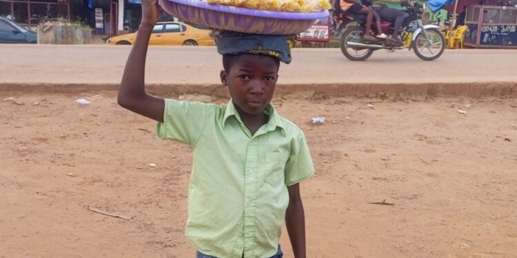 A young boy in Gbarnga, Bong County, carries a tray of snacks on his head while vending in the street instead of attending school.