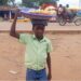 A young boy in Gbarnga, Bong County, carries a tray of snacks on his head while vending in the street instead of attending school.