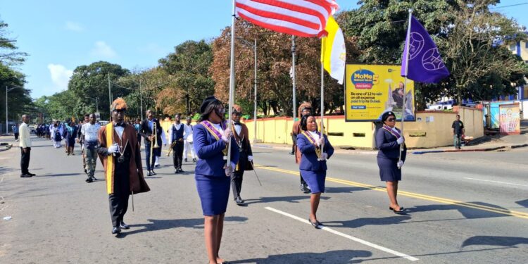 Ladies of Marshall members in ceremonial uniforms marching with the Liberian flag and organizational banner during a procession in Monrovia after Thanksgiving Mass.