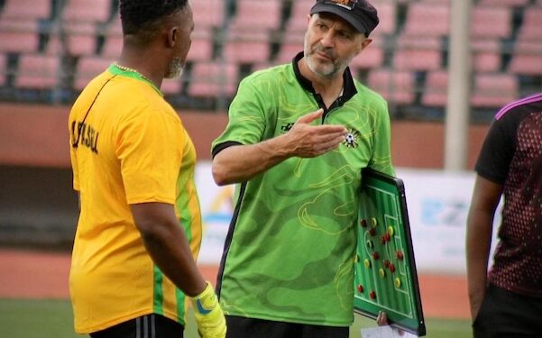 FC Fassell coach Amir Alagic strategizing with goalkeeper during a training session at the stadium, holding a tactics board while wearing the club’s green jersey.