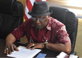 A man seated at a desk reviewing and signing official documents in an office setting.
