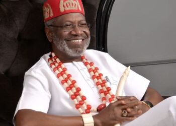 A man seated comfortably in traditional attire, wearing cultural regalia and smiling, with decorative beads and a carved staff visible
