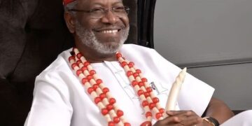 A man seated comfortably in traditional attire, wearing cultural regalia and smiling, with decorative beads and a carved staff visible