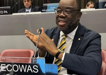 A speaker seated at a regional parliamentary meeting table, gesturing while addressing participants, with a conference nameplate in front