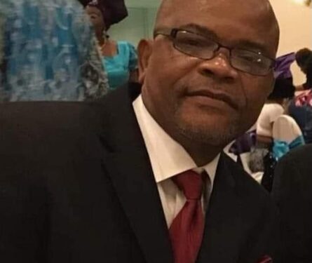 A man wearing a suit and glasses seated indoors during a formal gathering