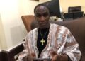 A man seated indoors wearing traditional attire and holding a mobile phone during an interview