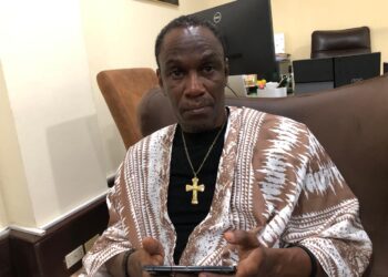 A man seated indoors wearing traditional attire and holding a mobile phone during an interview