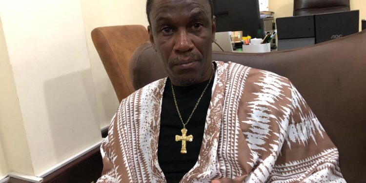 A man seated indoors wearing traditional attire and holding a mobile phone during an interview
