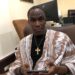 A man seated indoors wearing traditional attire and holding a mobile phone during an interview
