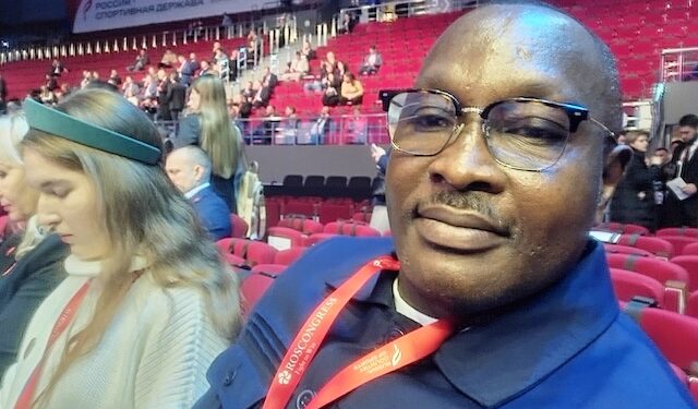 A man seated in an auditorium wearing glasses and a conference badge, with other attendees in the background.