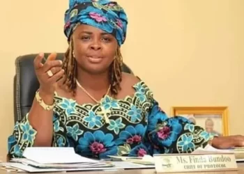 A woman in traditional attire seated at a desk, speaking and gesturing with documents in front of her.