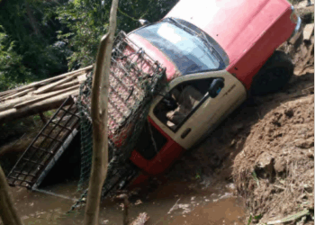 A pickup truck shown partially fallen or damaged on a deteriorated wooden bridge in a rural area, drawing attention to poor transportation infrastructure