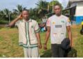 Two men standing outdoors in a rural community setting, posing for a photograph during daylight with homes and palm trees visible in the background.