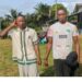 Two men standing outdoors in a rural community setting, posing for a photograph during daylight with homes and palm trees visible in the background.