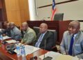 A panel of officials seated at a conference or legislative hearing table, with microphones and documents arranged in front of them.