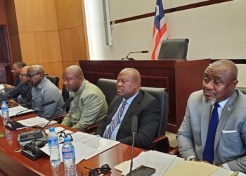 A panel of officials seated at a conference or legislative hearing table, with microphones and documents arranged in front of them.
