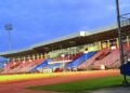 A football player in a red and blue kit posing on a pitch with a stadium structure in the background.
