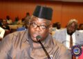 A man speaking into a microphone during a legislative hearing, seated among other participants in a conference hall