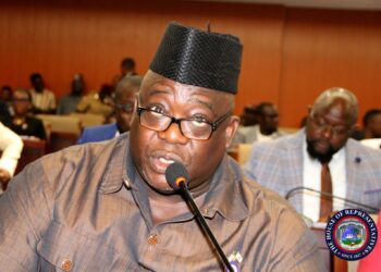 A man speaking into a microphone during a legislative hearing, seated among other participants in a conference hall