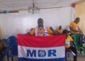 A man standing indoors holding a red, white, and blue MDR party flag, with others seated behind him.