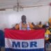 A man standing indoors holding a red, white, and blue MDR party flag, with others seated behind him.