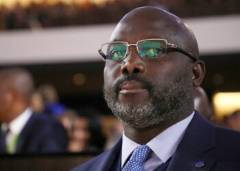 A man in a suit wearing glasses, seated at a formal event, looking forward with a calm expression.