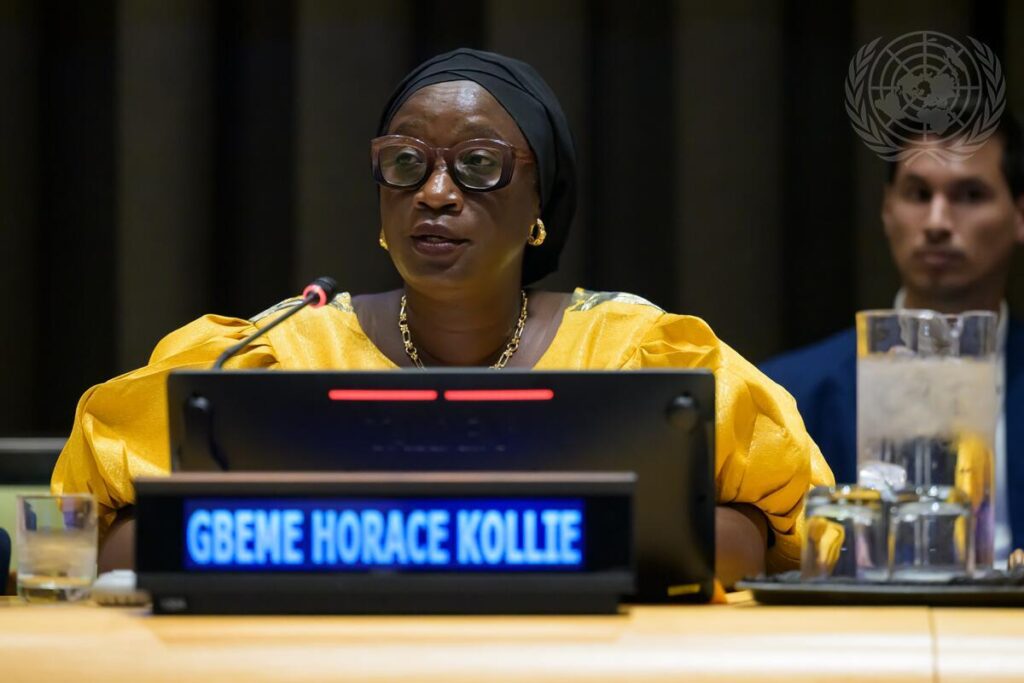 A woman in formal attire speaking at a panel in a conference setting, seated behind a microphone and nameplate, with other attendees visible in the background.