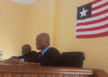 A judge seated at a courtroom bench with another official beside him, with a national flag displayed on the wall behind them.