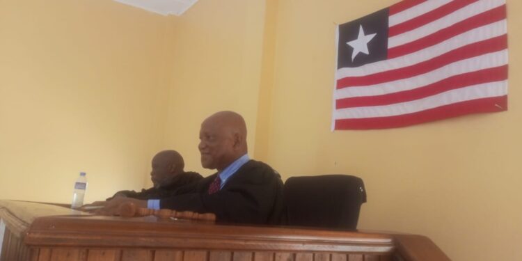 A judge seated at a courtroom bench with another official beside him, with a national flag displayed on the wall behind them.