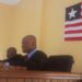 A judge seated at a courtroom bench with another official beside him, with a national flag displayed on the wall behind them.