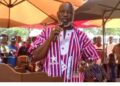 A man speaking into a microphone at an outdoor public event, wearing patterned traditional attire, with supporters seated behind him