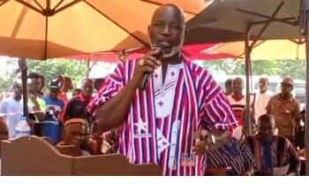 A man speaking into a microphone at an outdoor public event, wearing patterned traditional attire, with supporters seated behind him