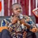 Jefferson T. Koijee speaking into a microphone during a public forum, appearing serious as he addresses political concerns in Liberia