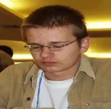 International Master Štefan Mačák seated indoors, wearing glasses and focusing on a chess activity during a tournament or training session.