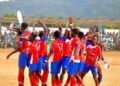Players in red and blue county jerseys celebrating on a dirt football pitch during a National County Meet match as spectators look on from the stands.