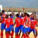 Players in red and blue county jerseys celebrating on a dirt football pitch during a National County Meet match as spectators look on from the stands.