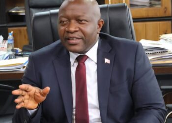 A man in a dark suit and red tie seated behind a desk, gesturing with his hand while speaking in an office settin