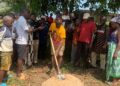 Representative Luther Sandy Collins performs groundbreaking with a shovel as community members gather to launch the maternity waiting home project at Kpayeakwelleh Clinic.