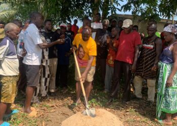 Representative Luther Sandy Collins performs groundbreaking with a shovel as community members gather to launch the maternity waiting home project at Kpayeakwelleh Clinic.