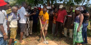 Representative Luther Sandy Collins performs groundbreaking with a shovel as community members gather to launch the maternity waiting home project at Kpayeakwelleh Clinic.