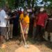 Representative Luther Sandy Collins performs groundbreaking with a shovel as community members gather to launch the maternity waiting home project at Kpayeakwelleh Clinic.