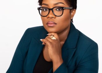 Cornelia Kruah wearing glasses and a teal suit seated against a plain background