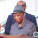 President Joseph Boakai speaking at a podium wearing a gray suit and hat during an official proclamation ceremony