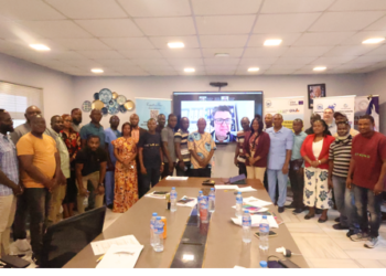 Group of workshop participants standing around a conference table during a fisheries management meeting in Monrovia with a virtual speaker displayed on a screen