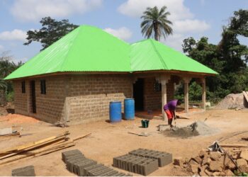 A beneficiary stands beside a house built with proceeds from the Salayea Community Forest village savings and loan scheme in Lofa County.