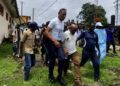 Police escort Rep. Thomas Romeo Quioh through the University of Liberia’s Capitol Hill campus as students gather nearby